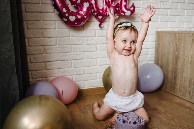 Baby raising hands in excitement during a Cake Smash Photoshoot with birthday cake and balloons.