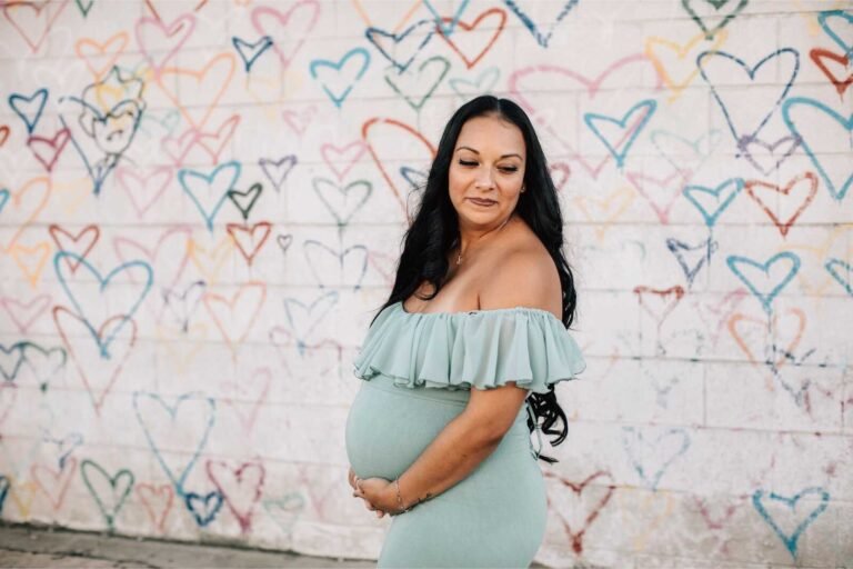 Pregnant woman in a soft off-shoulder gown posing by a colorful wall as inspiration for What to Wear to a Maternity Photo Shoot.