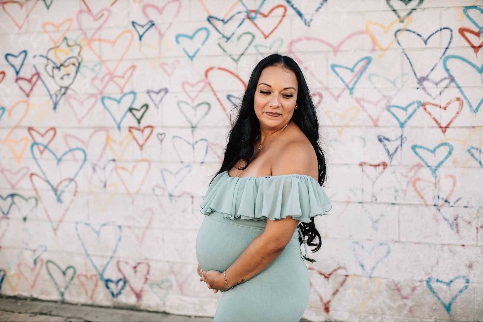 Pregnant woman in a soft off-shoulder gown posing by a colorful wall as inspiration for What to Wear to a Maternity Photo Shoot.
