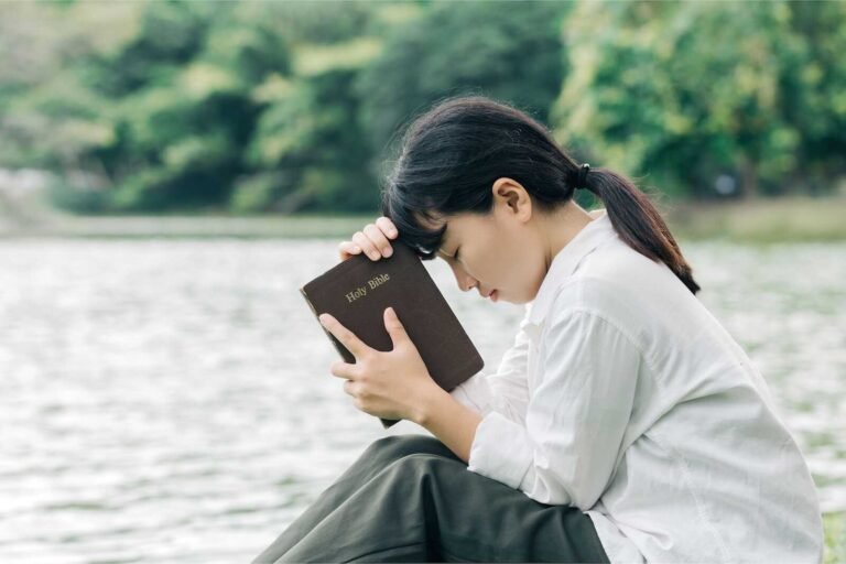 Woman holding a Bible and praying by a lake, symbolizing a heartfelt prayer for homeless individuals.