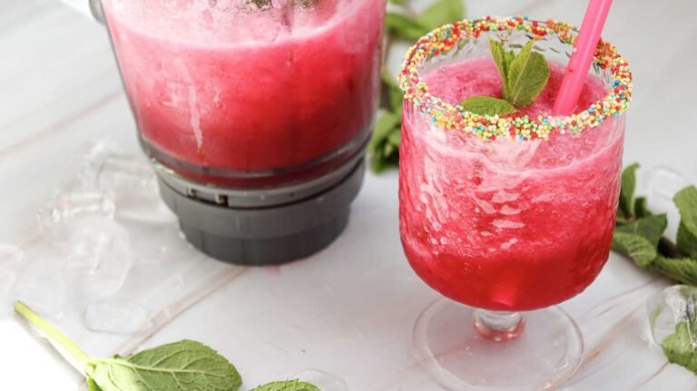 Watermelon slushie in sprinkle-rimmed glass with blender in background.