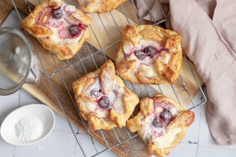 Golden Danish pastries with cream cheese and cherries dusted with powdered sugar on a cooling rack.