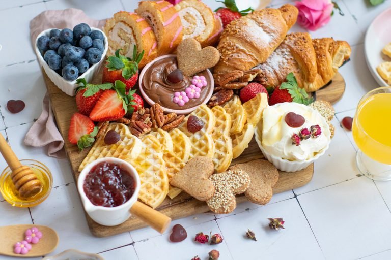 Sweet breakfast board with heart waffles, croissants, fresh berries, cookies, pecans, and sweet spreads.